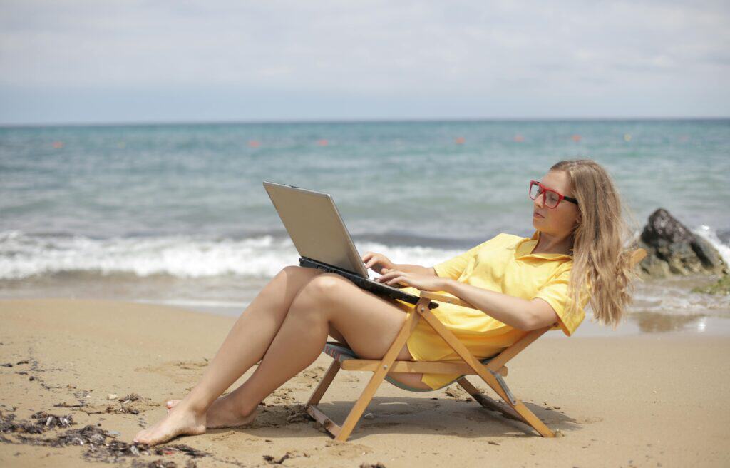 A woman with blond hair wearing a yellow top relaxes on a beach chair while using a laptop by the seashore.